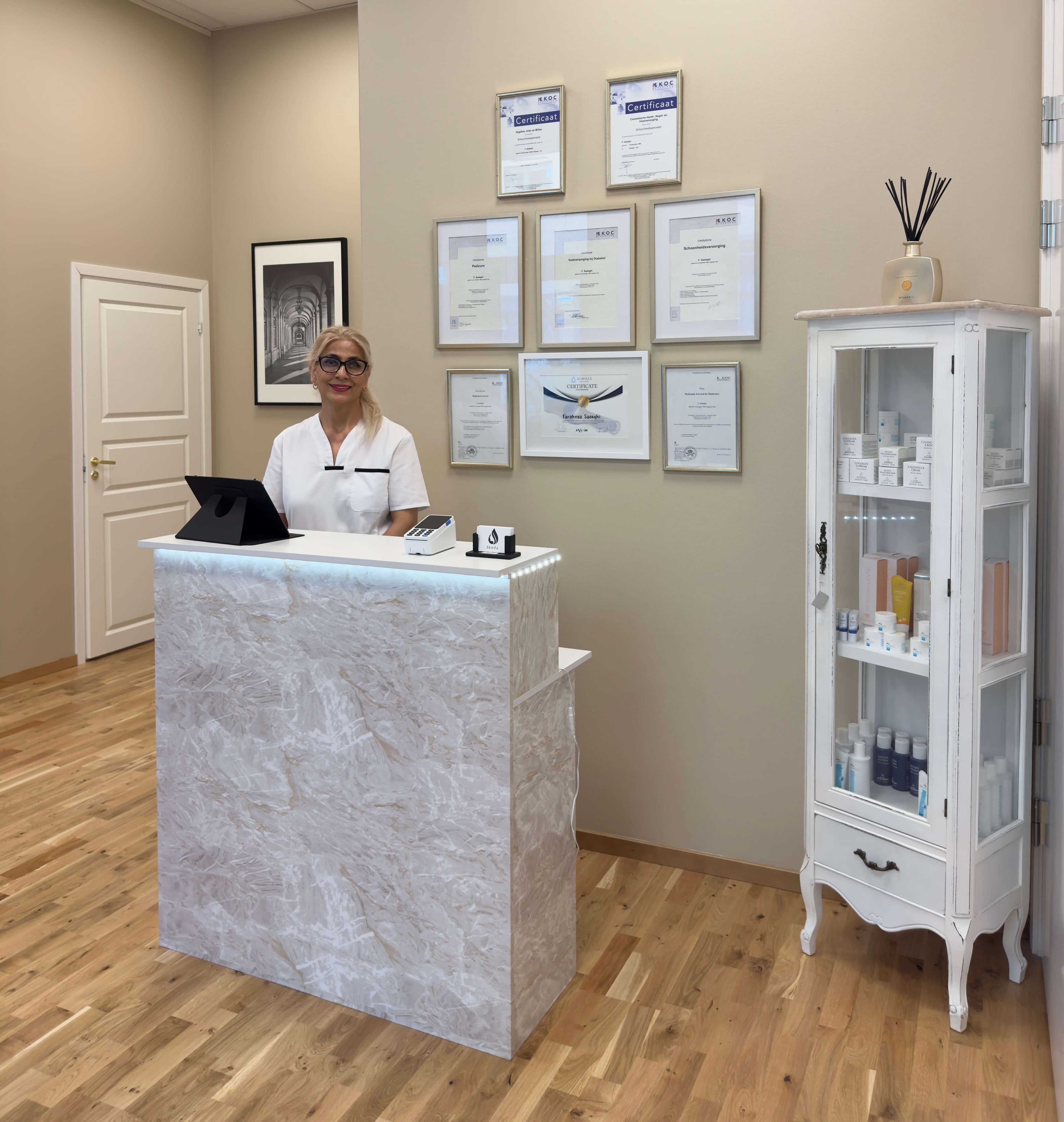 Hudvårdsspecialist vid receptionen på SkinFa salong i Stockholm, omgiven av diplomer och hudvårdsprodukter – professionell och certifierad skönhetsklinik med fokus på ansiktsbehandlingar och medicinsk fotvård.

